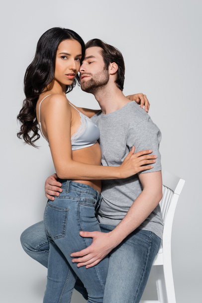 sexy woman in jeans and bra looking at camera while seducing man sitting on armchair on grey - Zdjęcie, obraz