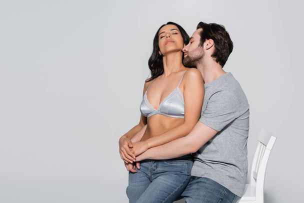 man in t-shirt hugging passionate woman in satin bra while sitting on chair on grey - Photo, Image