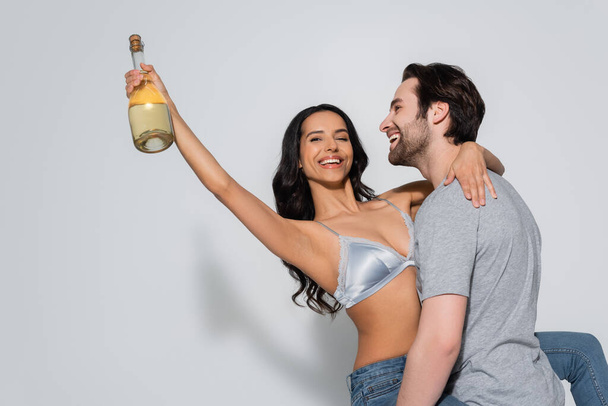 happy man in t-shirt holding sexy woman with champagne bottle on grey - 写真・画像