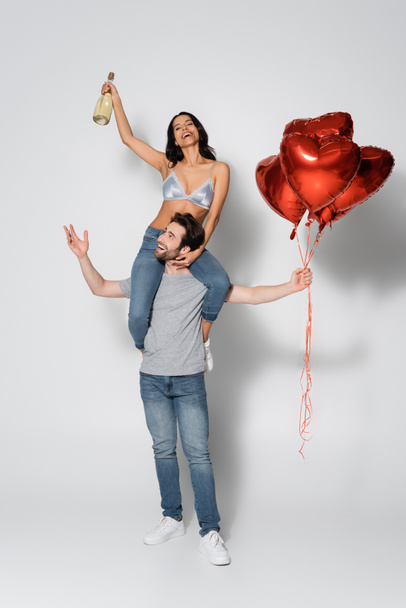 feliz hombre con globos rojos piggybacking mujer en sujetador celebración botella de champán en gris - Foto, Imagen
