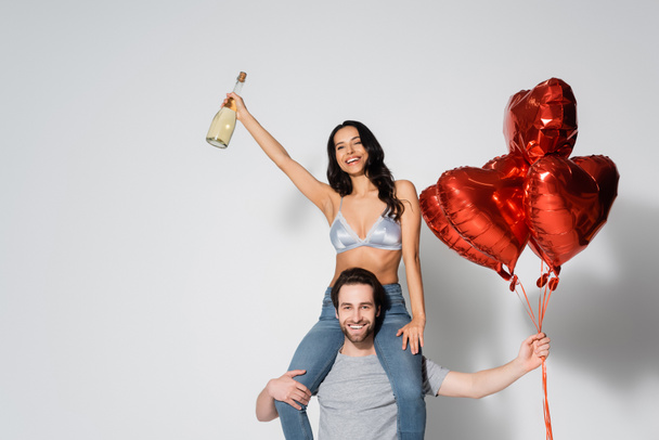άνδρας με μπαλόνια σε σχήμα καρδιάς piggybacking σέξι και ευτυχισμένη γυναίκα με μπουκάλι σαμπάνιας σε γκρι - Φωτογραφία, εικόνα