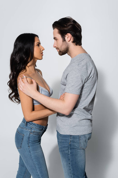 side view of brunette woman in bra embracing man in t-shirt on grey - Photo, Image