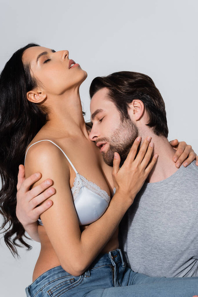 man with closed eyes embracing woman in satin bra isolated on grey - Фото, зображення
