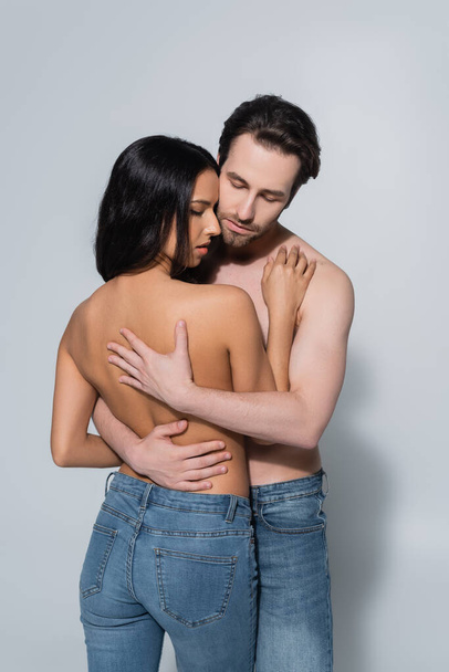 молодий без сорочки чоловік із закритими очима обіймає жінку в джинсах на сірому
 - Фото, зображення
