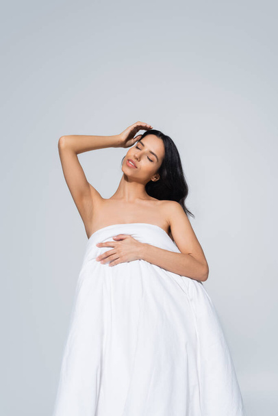 young woman with naked shoulders and closed eyes posing with white blanket isolated on grey - Fotografie, Obrázek
