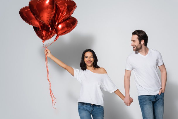mujer alegre con globos en forma de corazón tomados de la mano con el novio joven en gris - Foto, imagen