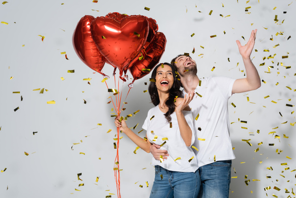 cheerful couple with heart-shaped balloons laughing under falling confetti on grey - 写真・画像