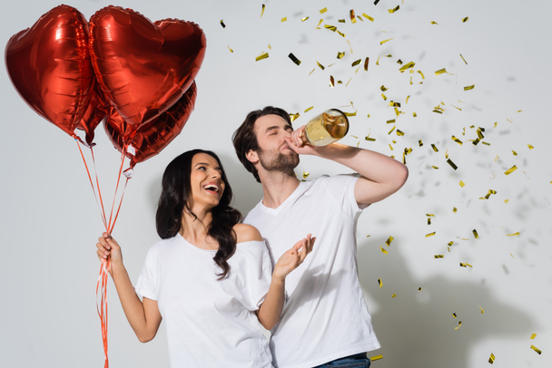 vzrušená žena drží balónky ve tvaru srdce a muž pije šampaňské z láhve poblíž konfety na šedi - Fotografie, Obrázek