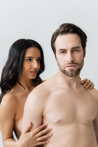 sexy brünette Frau umarmt hemdlosen Mann Blick auf Kamera isoliert auf grau - Foto, Bild