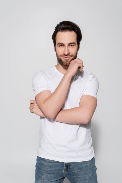 positive man in white t-shirt holding hand near face while looking at camera on grey - Fotografie, Obrázek