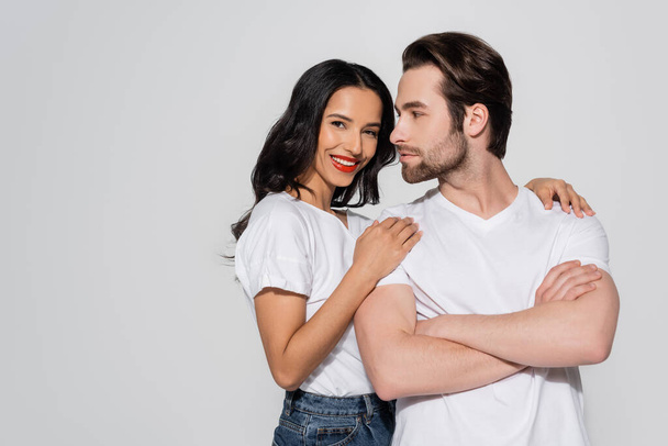 smiling brunette woman looking at camera while hugging shoulders of man standing with crossed arms isolated on grey - Foto, Bild