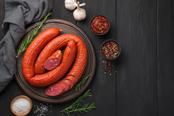 Rings of smoked Krakow sausage with sliced pieces on a black wooden background. Top view, copy space. - Photo, Image