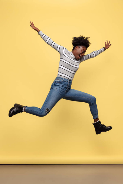 Full-length portrait of happy African girl in casual clothes jumping isolated on yellow studio background. Concept of emotions, holidays - Foto, imagen