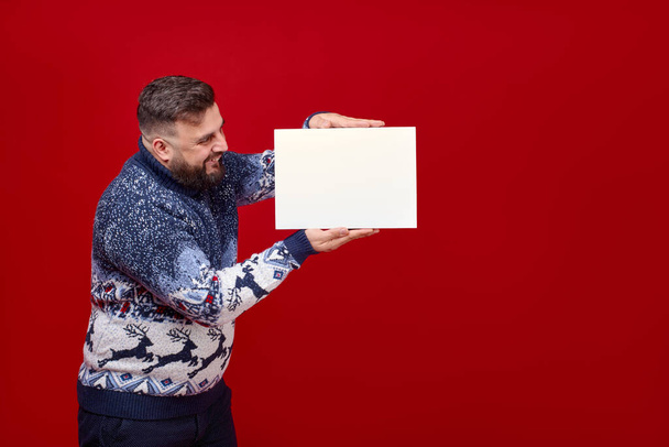 Een man in een oudejaarsweater heeft een doos op een rode achtergrond. - Foto, afbeelding
