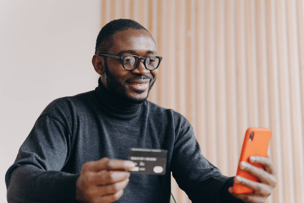 Młody, szczęśliwy afrykański przedsiębiorca z kartą kredytową za pomocą aplikacji e-banking na smartfonie - Zdjęcie, obraz