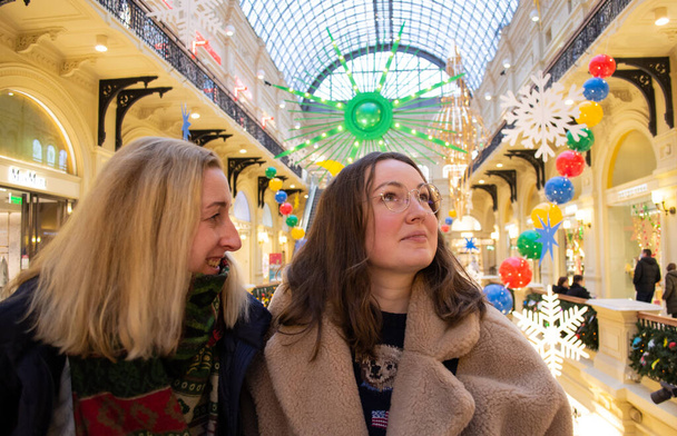 Deux jeunes belles amies au centre commercial souriantes, admirent le décor lumineux de Noël. - Photo, image