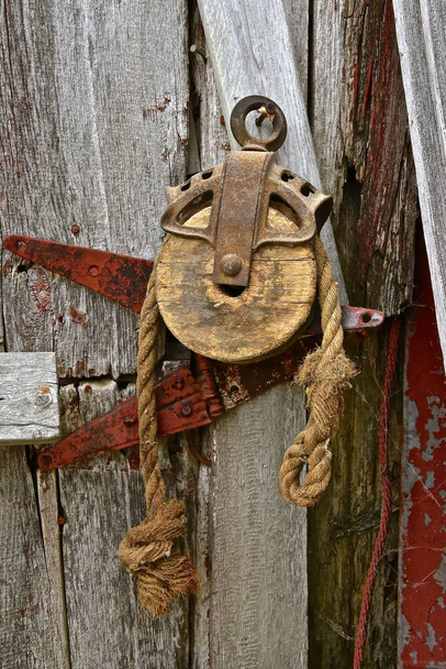 Старый деревянный шкив с концом изношенной веревки, висящей на выветренной двери сарая - Фото, изображение