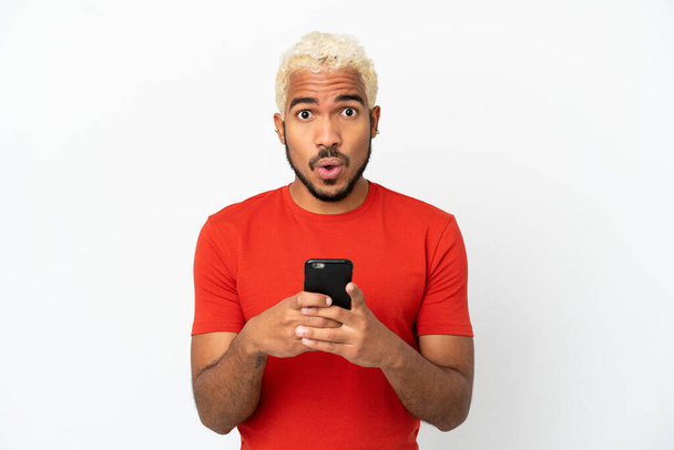 Young Colombian handsome man isolated on white background looking at the camera while using the mobile with surprised expression - Фото, изображение