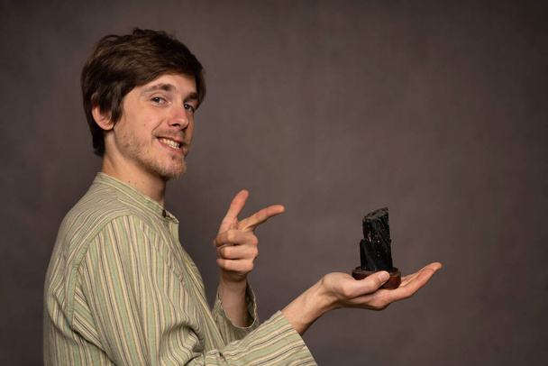 Young handsome tall slim white man with brown hair smiling and pointing at black crystal in striped shirt on grey background - Foto, Imagem