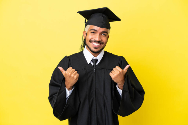 młody absolwent uniwersytetu Kolumbijczyk odizolowany na żółtym tle z kciukami w górze gest i uśmiech - Zdjęcie, obraz