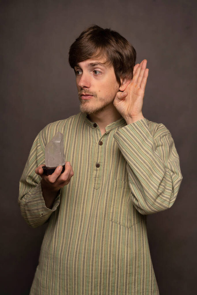 Young handsome tall slim white man with brown hair holding crystal trying to hear in striped shirt on grey background - Foto, imagen