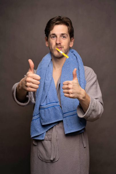 Young handsome tall slim white man with brown hair brushing his teeth thumbs up in grey bathrobe on grey background - 写真・画像