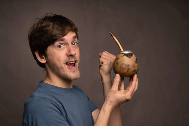 Young handsome tall slim white man with brown hair enjoying himself holding mate cup in grey shirt on grey background - Foto, Imagen