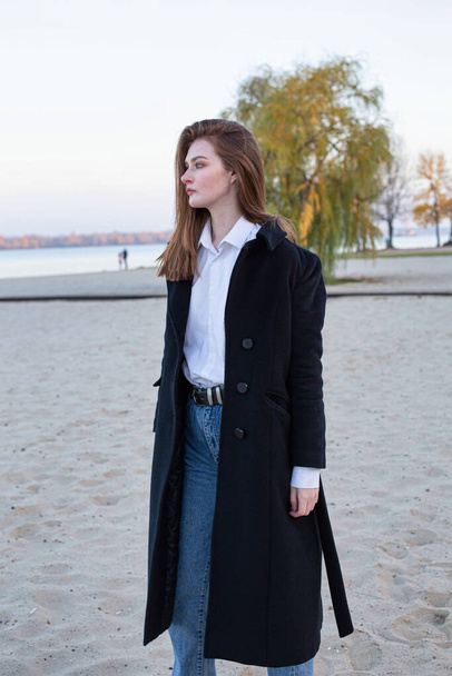 Long straight haired white european woman in a white shirt in dark coat in autumn on the beach - Фото, зображення