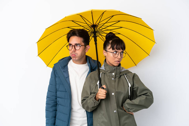 jong gemengd ras paar met een paraplu geïsoleerd op witte achtergrond met verwarde gezicht uitdrukking terwijl beten lip - Foto, afbeelding