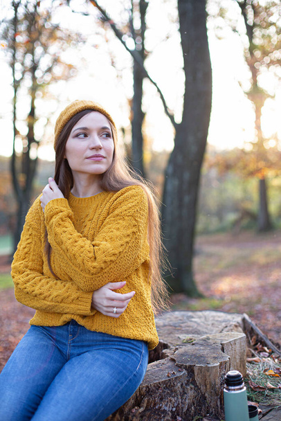 Young european white woman in yellow sweater in sunny autumn forest sitting on tree stump - Fotó, kép