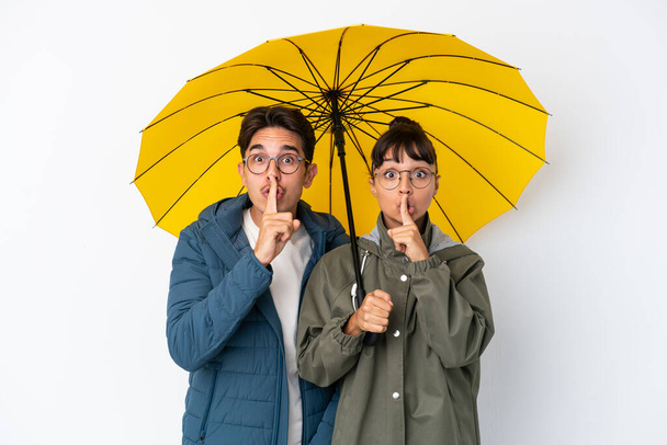 Nuori sekarotuinen pariskunta, jolla on valkoisella pohjalla eristetty sateenvarjo, jossa näkyy hiljaisuuden ele sormen laittamisesta suuhun - Valokuva, kuva