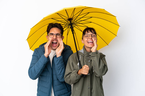 jong gemengd ras paar met een paraplu geïsoleerd op witte achtergrond schreeuwen en aankondigen iets - Foto, afbeelding