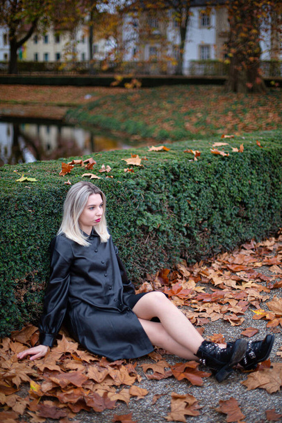 Young european white woman with blonde hair in total black sitting on the ground in autumn park - Foto, afbeelding