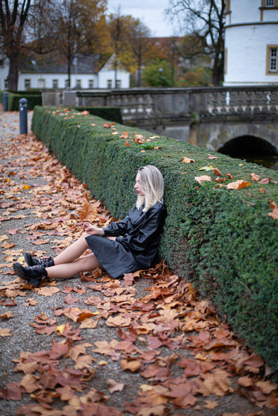 White young european woman with blonde hair in total black sitting on the ground in autumn park - Valokuva, kuva