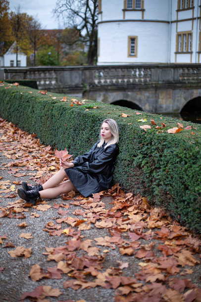 European white young woman with blonde hair in total black sitting on the ground in autumn park - Foto, immagini