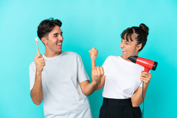 Young mixed race couple holding a hairdryer and toothbrush isolated on blue background celebrating a victory in winner position - Photo, Image