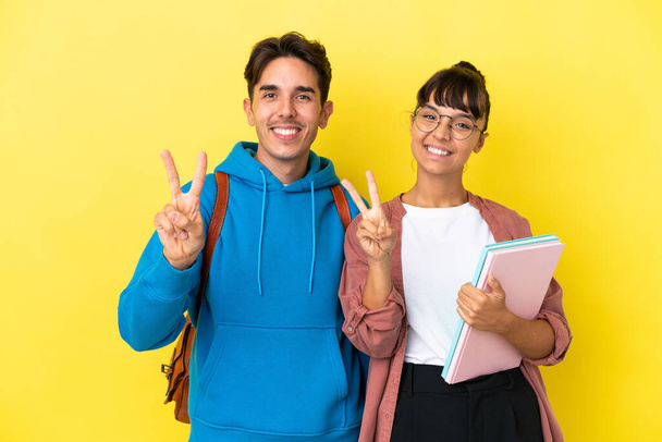 Young student couple isolated on yellow background smiling and showing victory sign - Foto, afbeelding