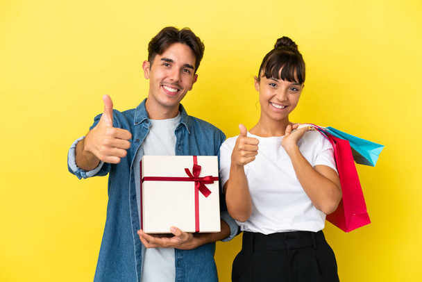 Young couple holding shopping bags and present isolated on yellow background giving a thumbs up gesture with both hands and smiling - Photo, Image