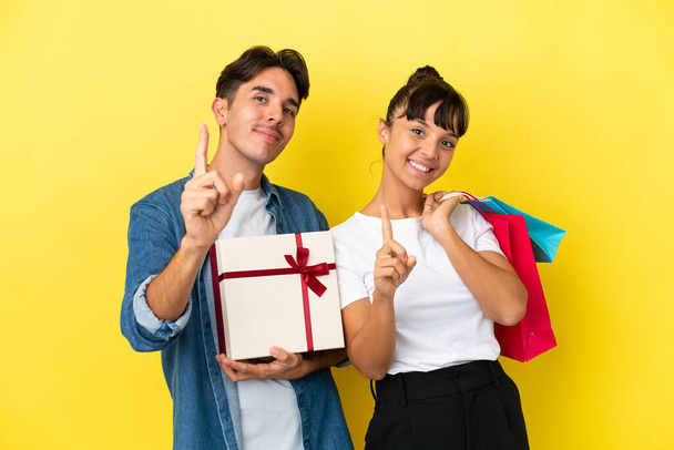 Giovane coppia che tiene borse della spesa e presente isolato su sfondo giallo che mostra e solleva un dito - Foto, immagini