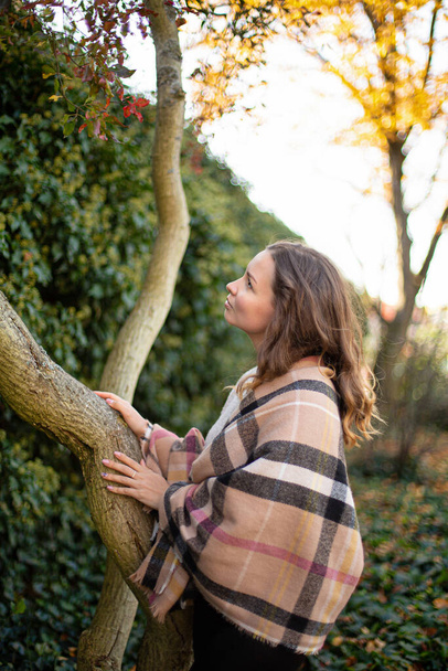 Looking up young european white woman with brown hair near tree trunk with bushes behind - Foto, afbeelding