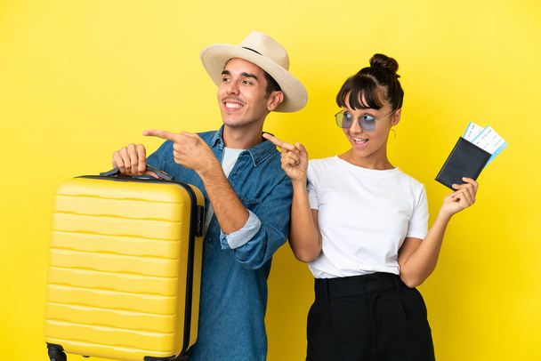 Jovens amigos viajantes segurando uma mala e passaporte isolado no fundo amarelo apontando o dedo para o lado em posição lateral - Foto, Imagem