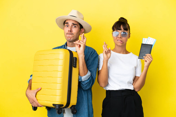 Jonge reizigers vrienden met een koffer en paspoort geïsoleerd op gele achtergrond met vingers kruisen en wensen de beste - Foto, afbeelding