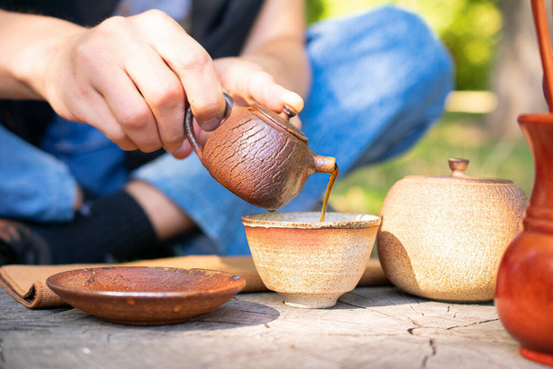 White man holding handmade ceramic tea pot with tea set on stump in city park - Zdjęcie, obraz