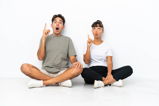 Jovem casal misto sentado no chão isolado no fundo branco pensando uma ideia apontando o dedo para cima - Foto, Imagem