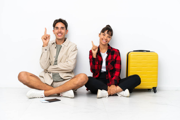 Молода пара мандрівників змішаної раси сидить на підлозі ізольовано на білому тлі, показуючи і піднімаючи палець на знак найкращих
 - Фото, зображення