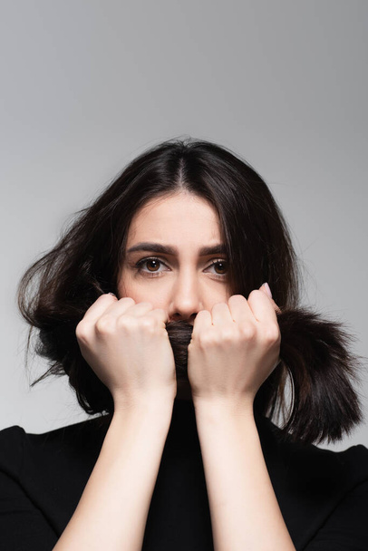 junge Frau bedeckt Mund mit glänzendem Haar isoliert auf grau - Foto, Bild