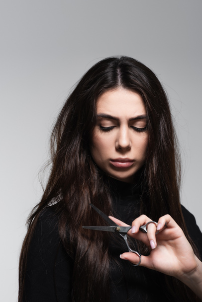young woman in black turtleneck holding scissors near long hair isolated on grey - Fotografie, Obrázek