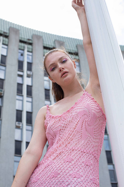 Portrait of white blond young woman with a ponytail in pink summer dress with blue eyes with building behind - Foto, Bild