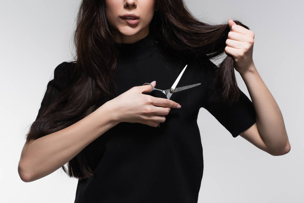частковий вигляд молодої жінки, що кусає губи і тримає ножиці біля довгого пошкодженого волосся ізольовані на сірому
 - Фото, зображення
