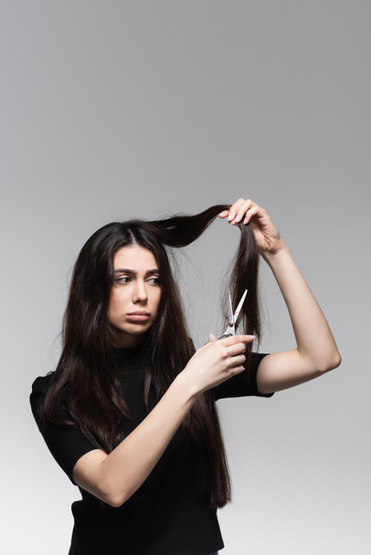 triste jovem mulher de gola alta preto segurando tesoura perto longo cabelo danificado isolado em cinza - Foto, Imagem